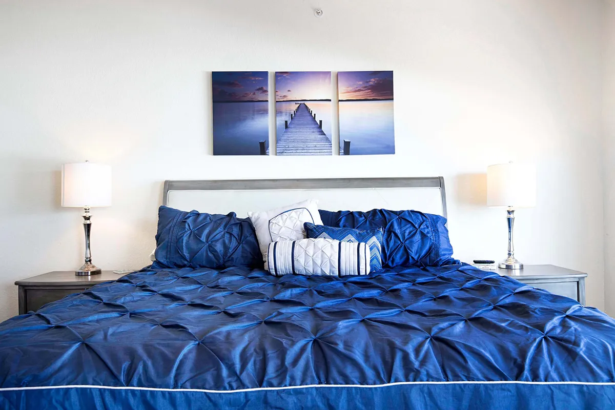 This image captures a serene bedroom scene with a bed adorned with decorative pillows and tasteful wall art. The overall setting is calm, bathed in soft light, and hints at the presence of scenic views—possibly from a nearby window—conveying a tranquil retreat ideal for couples seeking relaxation.