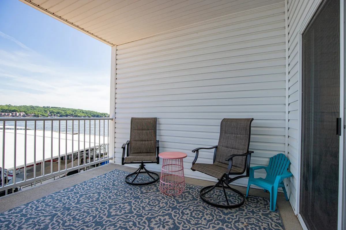 A cozy outdoor porch scene featuring two rocking chairs, a pink side table, a blue plastic chair, and an area rug, all oriented to take in the sunset. The arrangement invites relaxing, reading, and intimate conversations, blending comfort with the beauty of the lakeside environment.