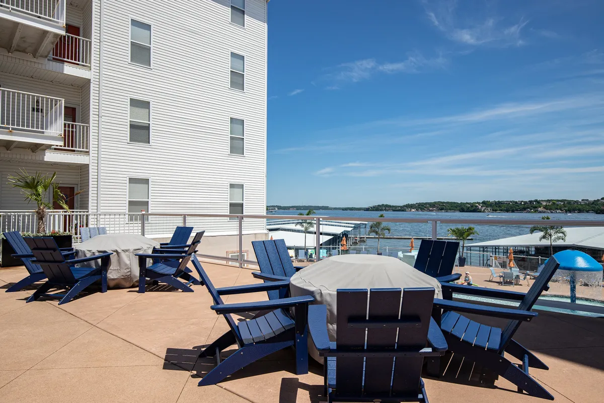 A waterfront patio scene featuring blue Adirondack chairs and a covered table, all overlooking the tranquil lake. The inviting setup highlights the close proximity to the water, and the seating arrangement suggests a perfect spot for morning coffee or evening relaxation with friends.