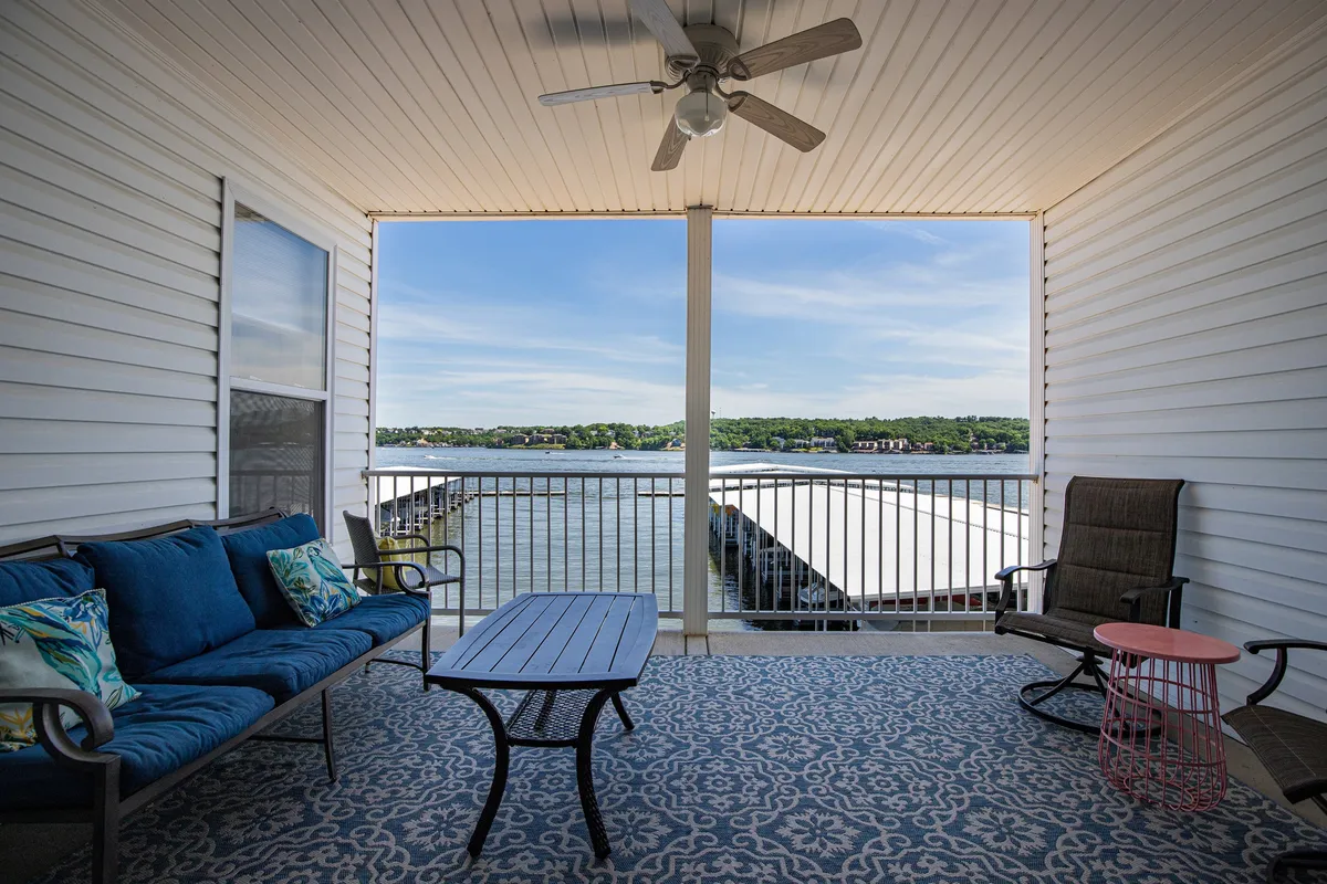 The image showcases a tranquil outdoor patio overlooking the shimmering Lake of the Ozarks, with a plush blue couch, a wooden table perfect for morning coffee or evening drinks, and a cozy rocking chair—all inviting guests to relax while soaking in the sweeping water views.