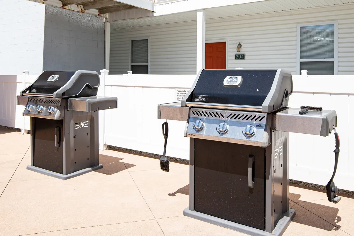 An outdoor grilling area with two charcoal grills, a white fence, and an orange door. The space is ideal for barbecues and gatherings, supporting a lifestyle filled with outdoor entertainment and community events.