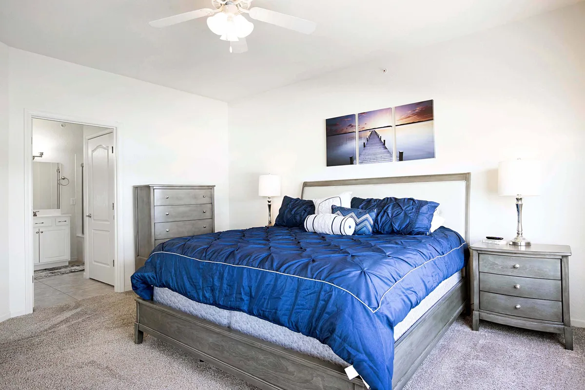 A serene bedroom with a large bed dressed in blue bedding, nightstands with lamps on each side, artwork above the bed, and a doorway leading to a bathroom. Soft lighting adds to the peaceful and comfortable atmosphere.