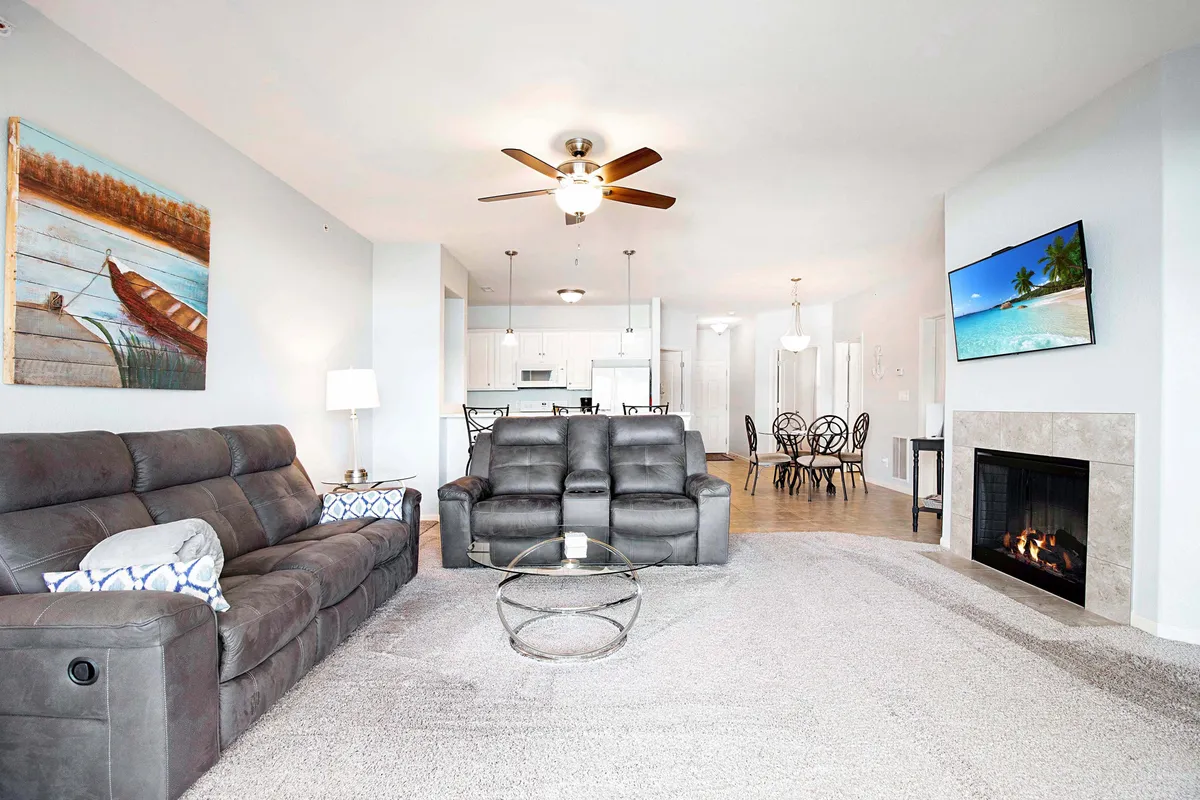 A wide view of the open-concept living and dining area, featuring comfortable gray sofas, decorative wall art, a TV mounted above an ornate electric fireplace, and a dining space with black chairs. Natural light floods in, and the inviting setup makes it perfect for relaxation or social gatherings.