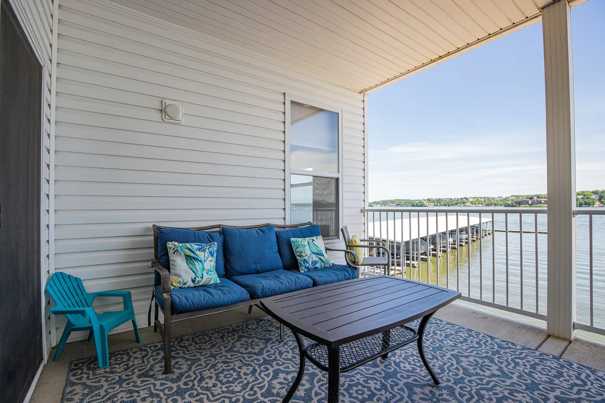 A serene balcony or patio setup featuring a blue couch with decorative pillows, a small turquoise chair, and a coffee table, all overlooking a sweeping water view. The scene evokes relaxation and the enjoyment of sunrise coffee or sunset gatherings, with the tranquil lake just beyond.
