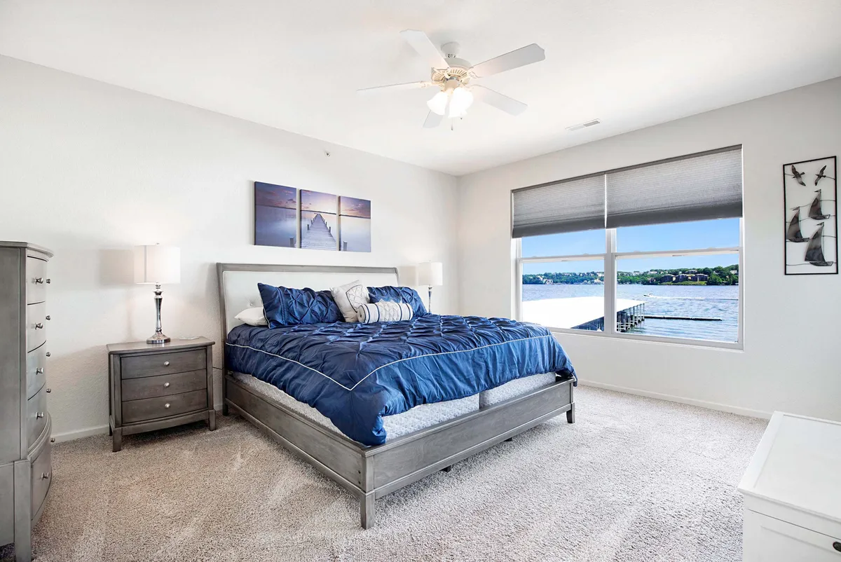 A tranquil bedroom featuring a bed with blue bedding, large window offering a scenic water view, and nightstands with lamps. Framed artwork adorns the wall, and natural light fills the space, enhancing its relaxing atmosphere.