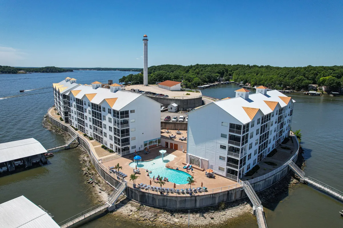 An exterior shot that captures the swimming pool, the architectural lighthouse, surrounding water, and building exteriors. The lively atmosphere hints at social activities like sunbathing, poolside barbecues, and fishing trips.