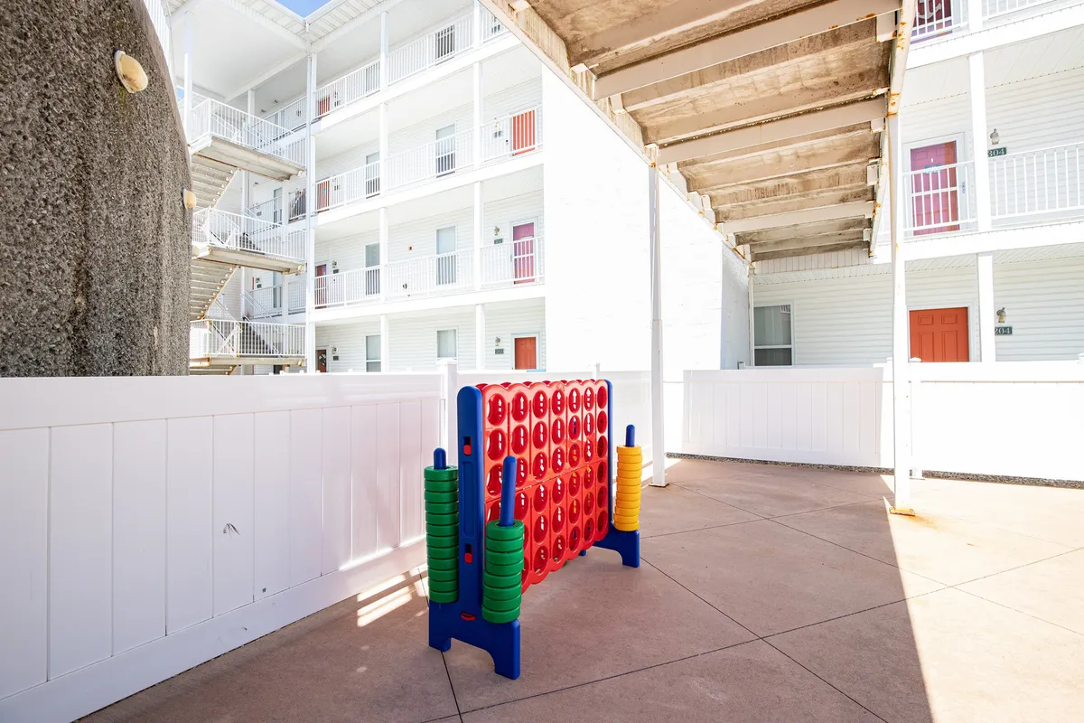 A lively outdoor space with a giant Connect Four game, white fence, and surrounding resort buildings. Children play and families gather, laughing and enjoying the playful environment.