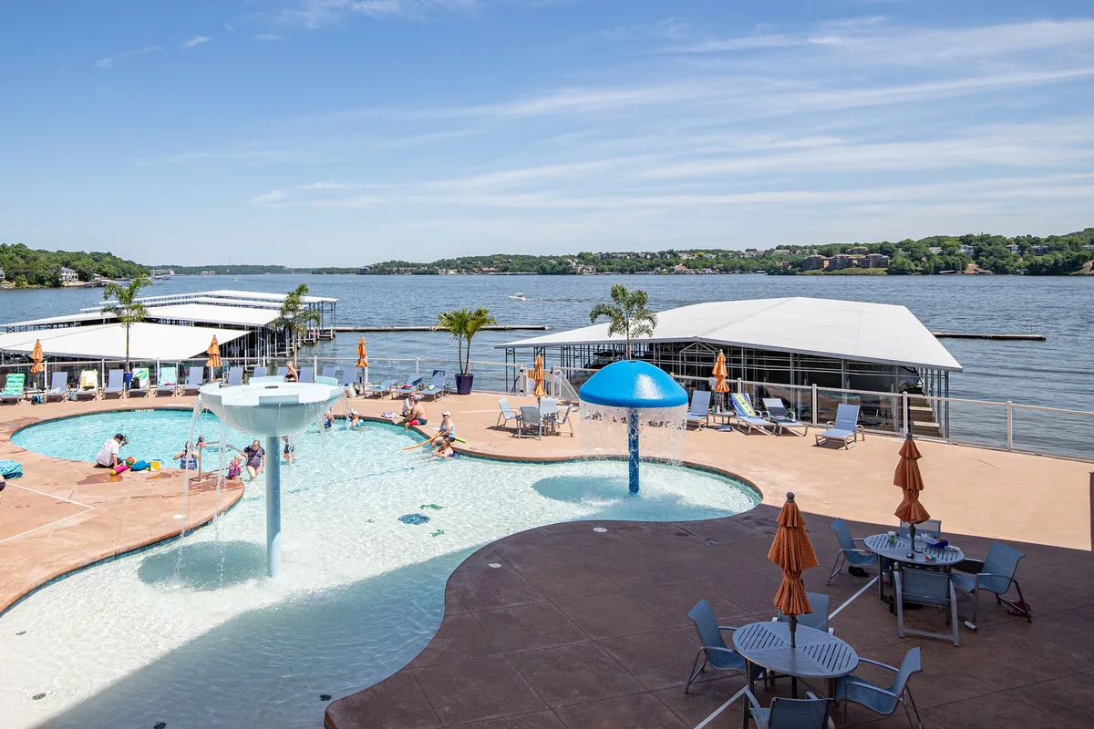 This exterior image features the lakeside zero-entry swimming pool with a prominent water fountain, blue mushroom umbrella, and an array of lounge chairs overlooking the lake. The inviting scene showcases the pool as a fun, social hub with the lake as a natural backdrop, emphasizing the family-friendly, resort-style atmosphere.